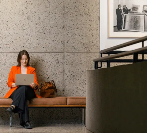 a person wearing an orange blazer sitting on a cushioned bench working on her laptop.