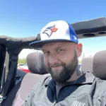 Picture of a caucasian man with mustache and beard, wearing a blue and white ball cap while sitting in an open-topped Jeep.