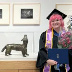 Picture of Jessica Colley, a young woman with pink hair, in a graduation cap and robe, holding flowes and a degree in a blue folder, standing next to an art gallery display of framed pictures and a sculpture.