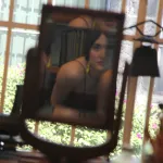 Photograph of a woman reflected, slightly out of focus, in a rectangular mirror in front of a bright, paned window.