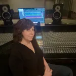 A photograph of Mary Mazurek from the waist up, sitting before a large control panel in a recording studio.  She is a Caucasian woman with dark, shoulder-length hair.