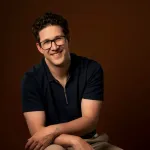 Picture of Alex Bohn, a caucasian man with short, dark hair and glasses, wearing a dark shirt, seated with forearms crossed in front of a brown background.