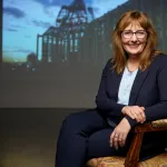 A photograph of Anne Dymond - a smiling woman with long, light brown hair, glasses, and a dark blue suit.  She is seated on an upholstered wooden chair, in a dark room with a slightly blurry architectural image projected on the wall behind her.