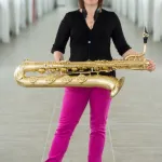 A picture of Holly De Caigny - a cauciasian woman with mid-length brown hair, wearing a black shirt and pink pants, holding a baritone saxophone, standing in a long hallway.