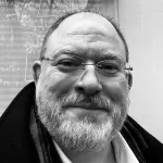 A black and white photograph of Dr. Arlan Schultz from the neck up, in front of a reflective window.  He is a caucasian man with short hair, a mustache, and beard, and is wearing rimless eyeglasses and a dark jacket.