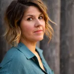 A photograph of Nicola Elson - a caucasian woman with mid-length hair, dark at the roots and lighter brown at the ends, pictured from the chest up in 3/4 profile.  She is wearting a blue blouse, and is standing in front of an unfocused, pillar-like bakground.