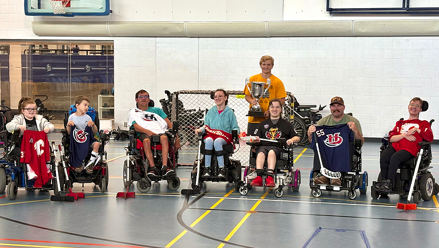 Powerchair hockey players display their jerseys while coach Chase Petruska holds the trophy.